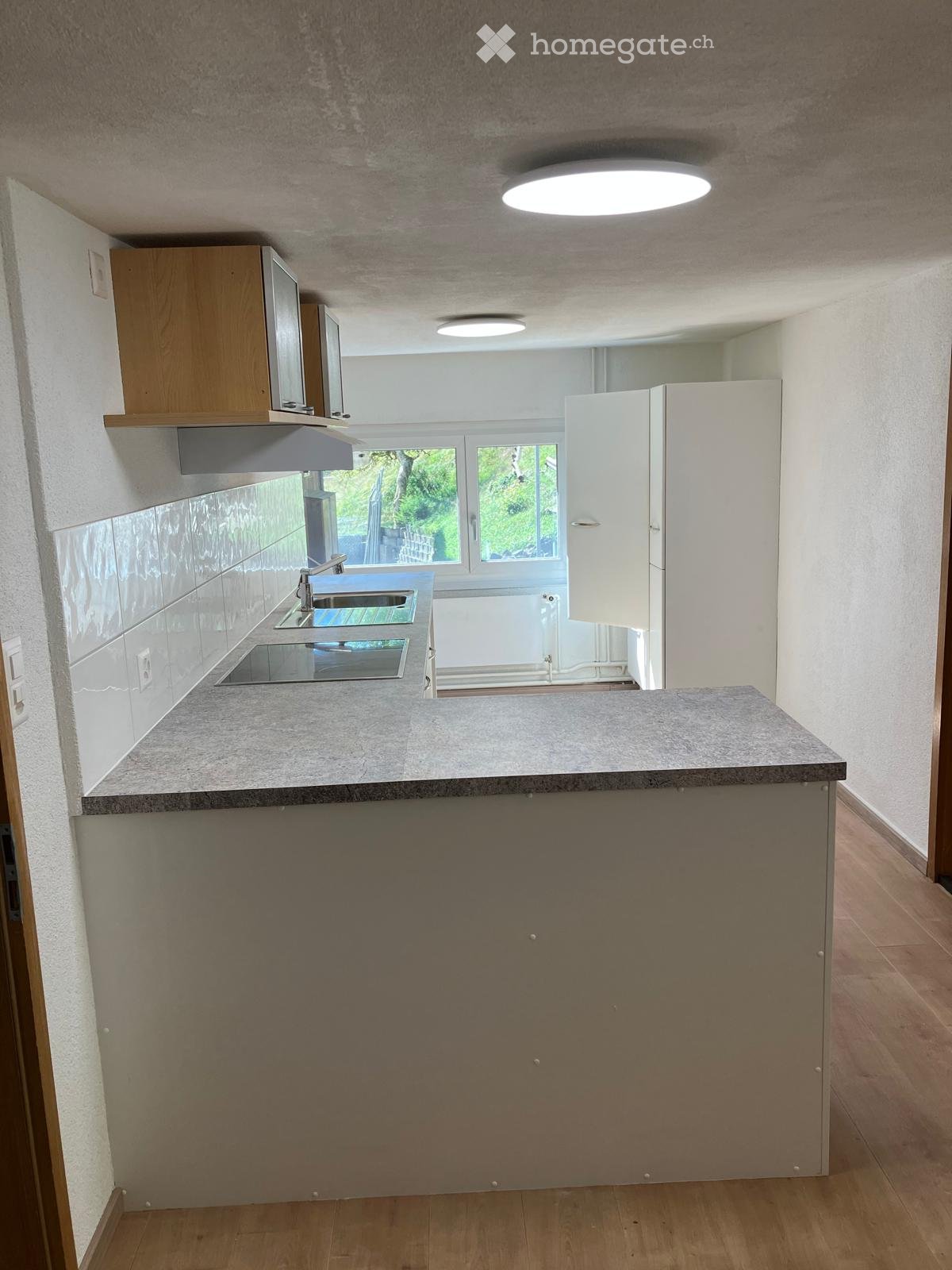 Modern kitchen with a grey countertop, built-in sink, wooden cabinets, and large windows offering a view of greenery.