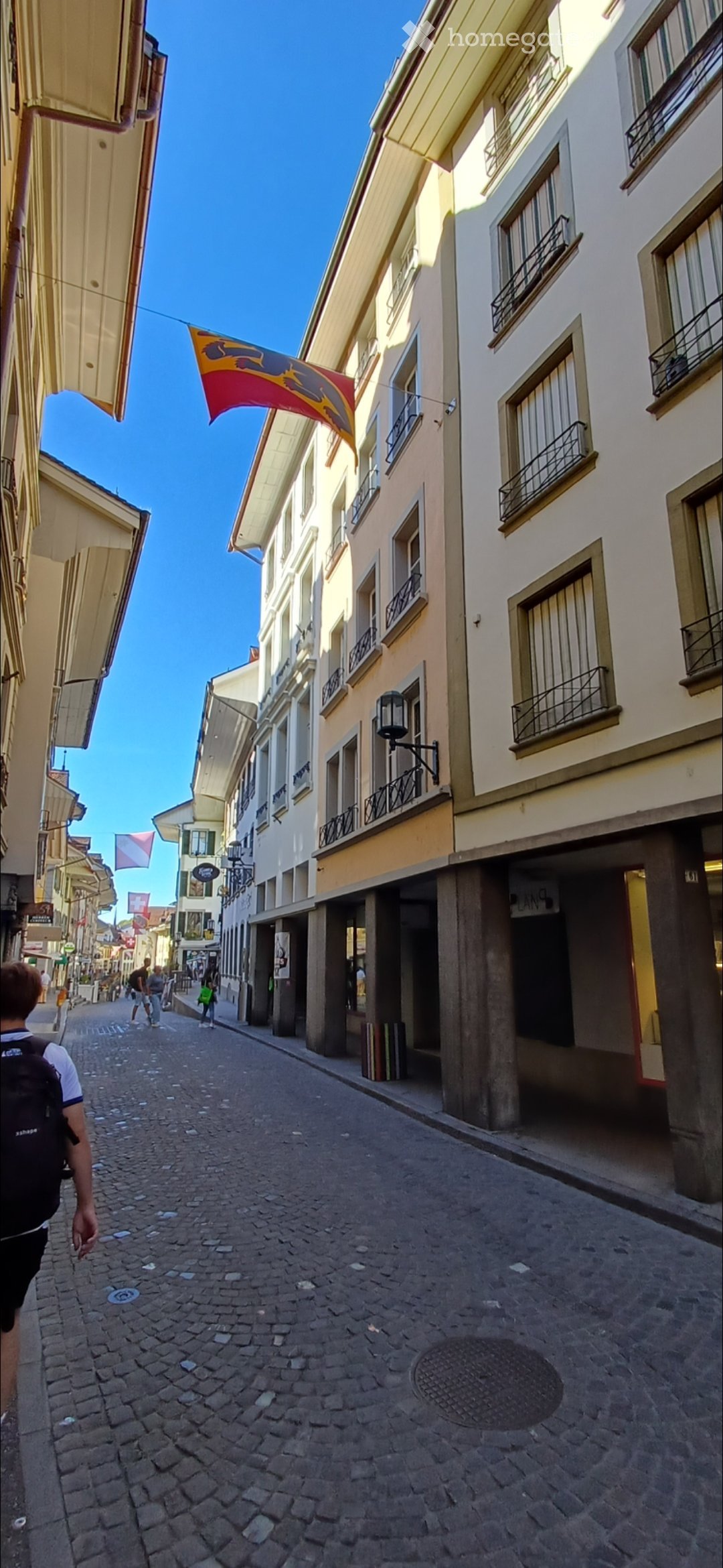A narrow cobblestone street lined with multi-story buildings featuring balconies and awnings. A flag is visible above the street.
