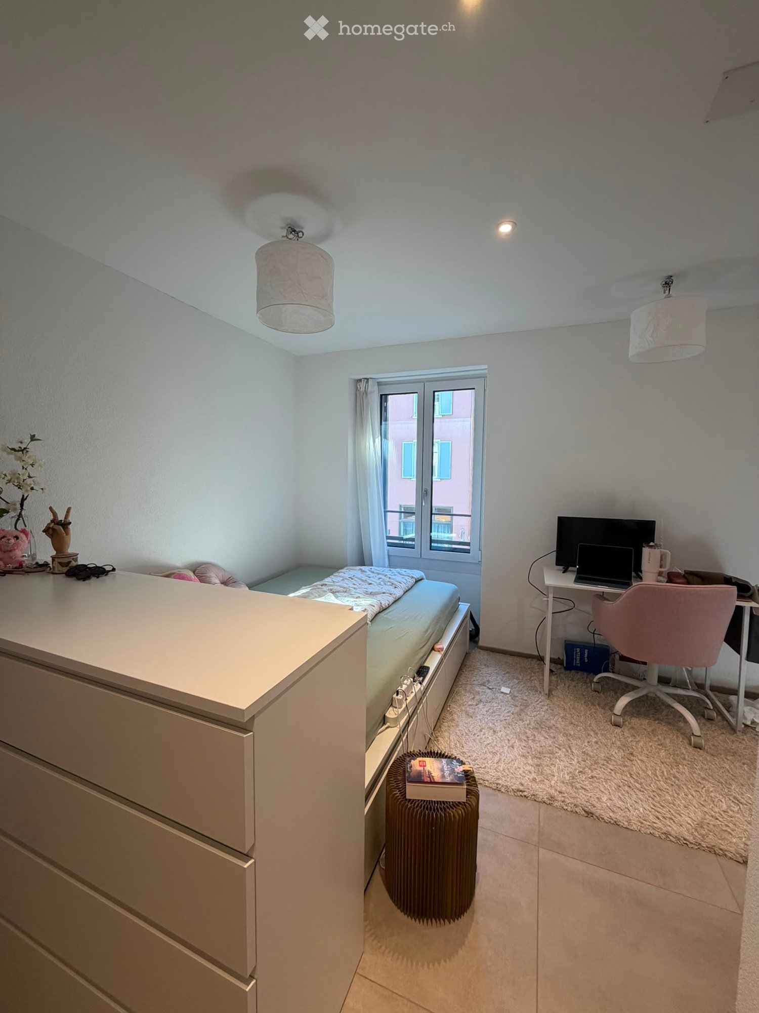 A well-lit bedroom with a bed, desk, chair, laptop, and a radiator. The room features a window with a view, a small shelf with decorative items, and a cozy carpeted area.