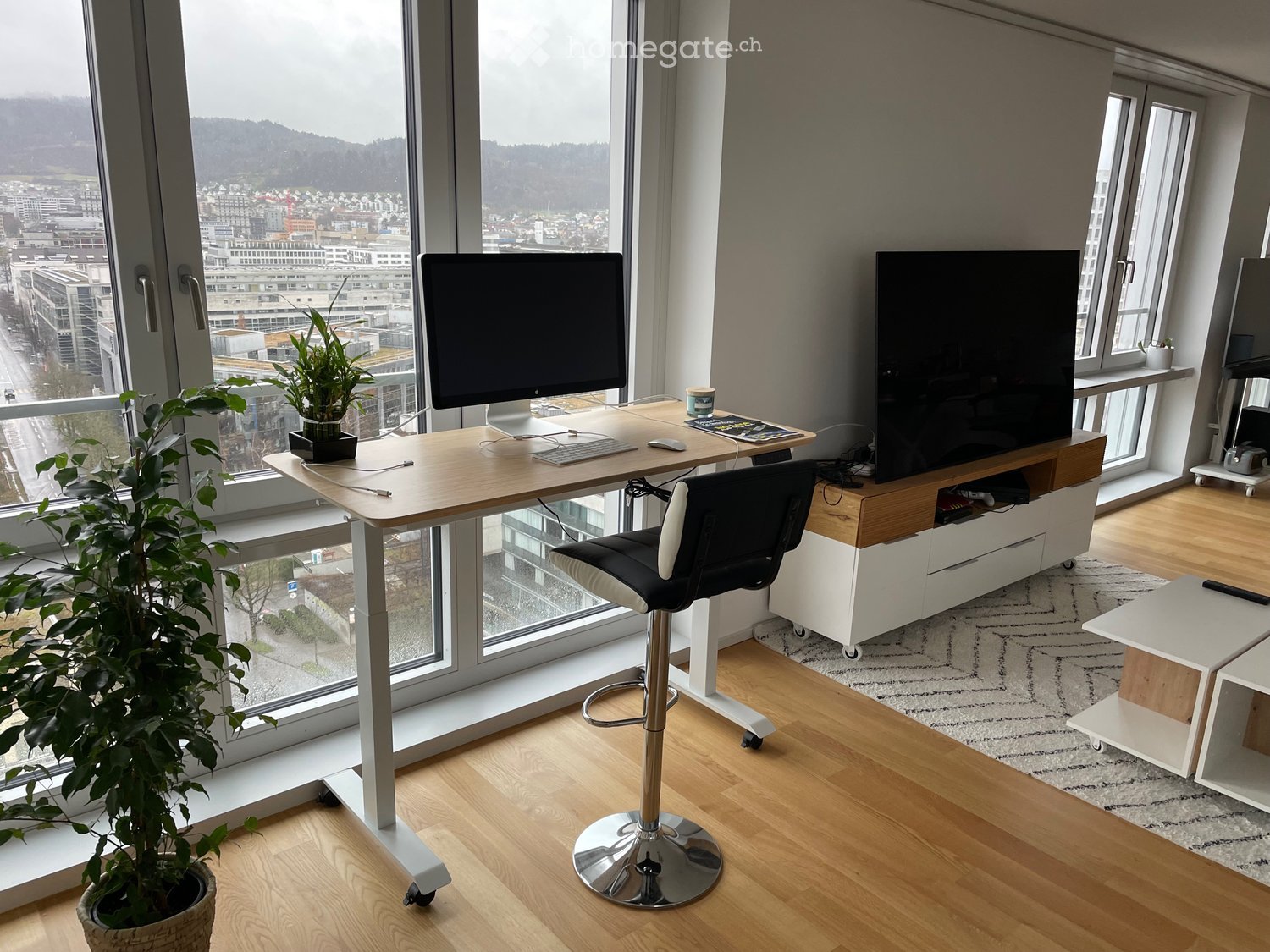 Modern, well-lit room with large windows offering a city view. Features a standing desk with a computer setup, a comfortable chair, a TV stand, a small table, and a potted plant.