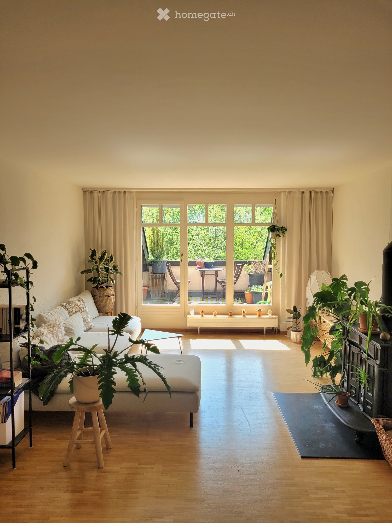 Bright living room with large windows and a balcony, featuring a white sofa, coffee table, potted plants, and a bookshelf.