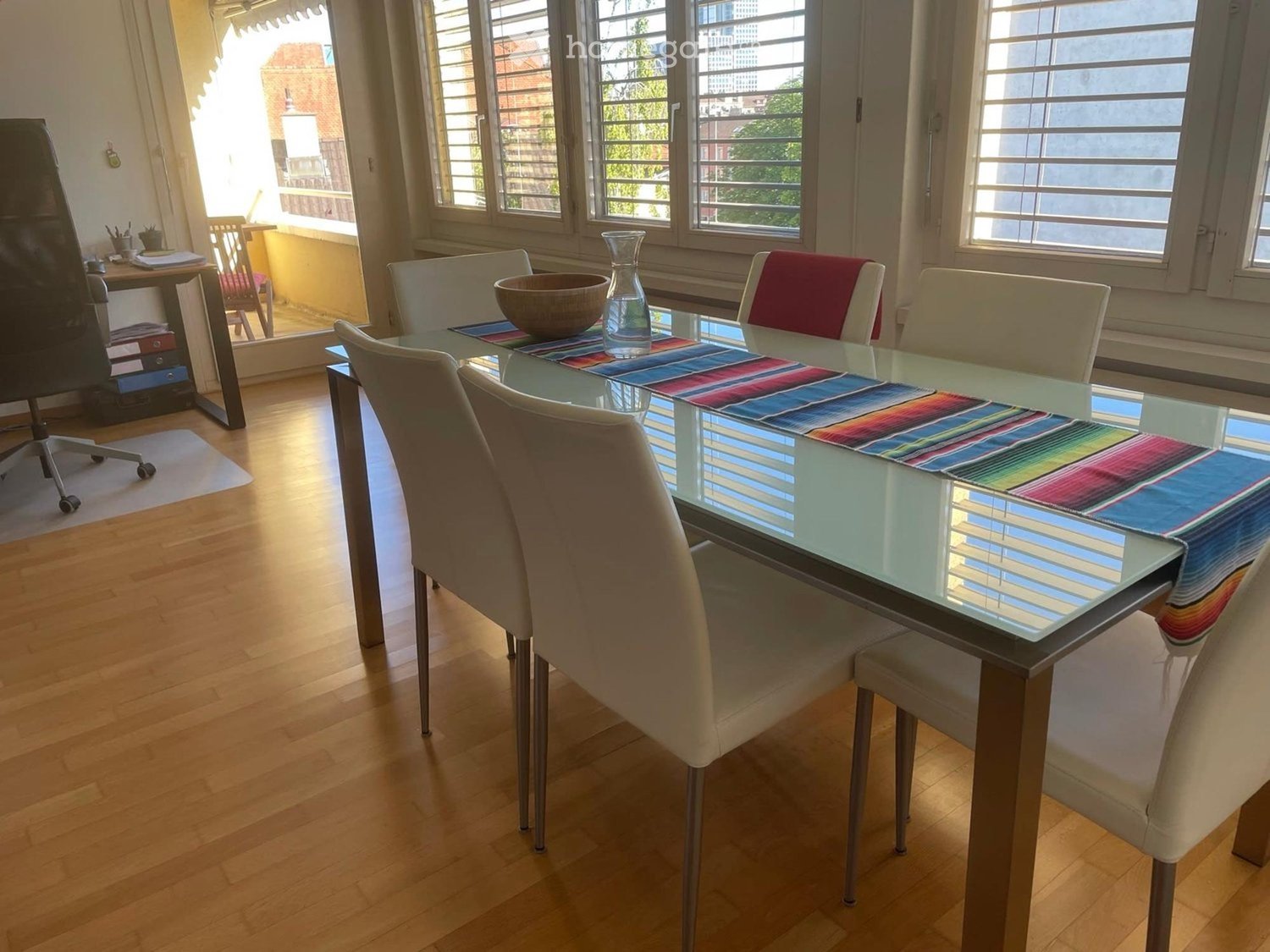Dining area with a glass table, colorful runner, six chairs, vase, bowl, and an office chair near a desk.