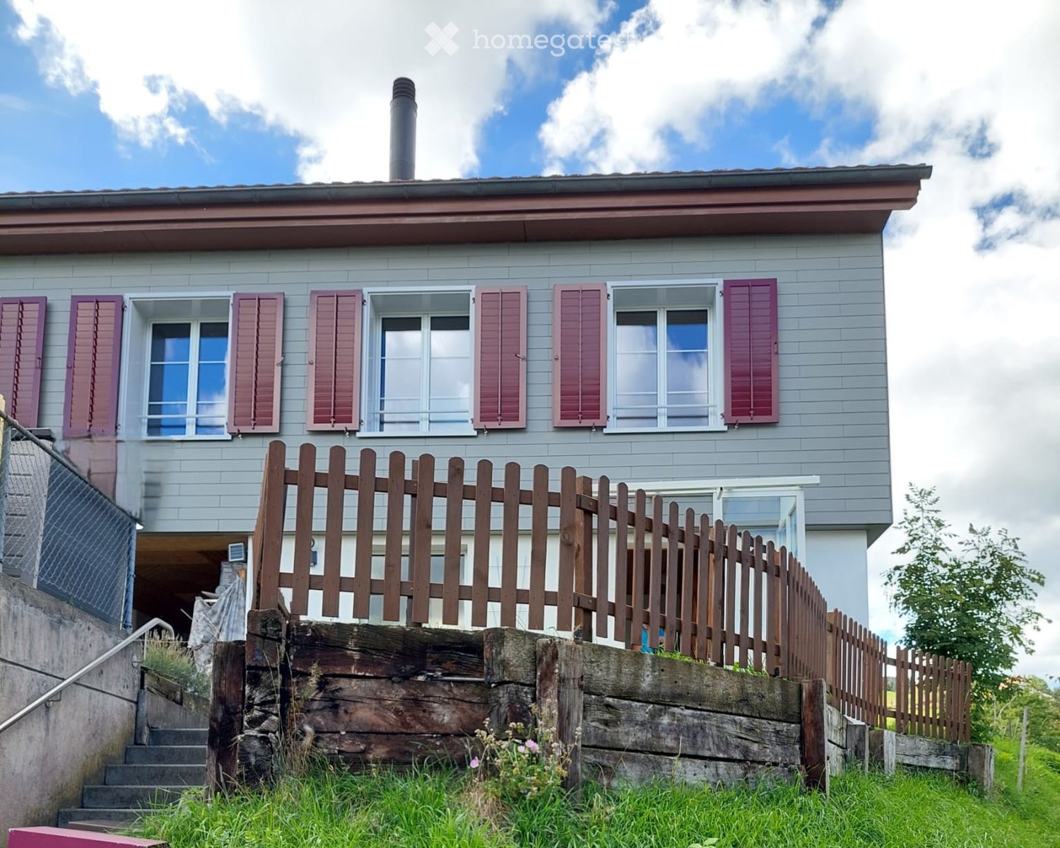 Two-story house with grey siding, multiple windows with shutters, and a wooden fence in front.