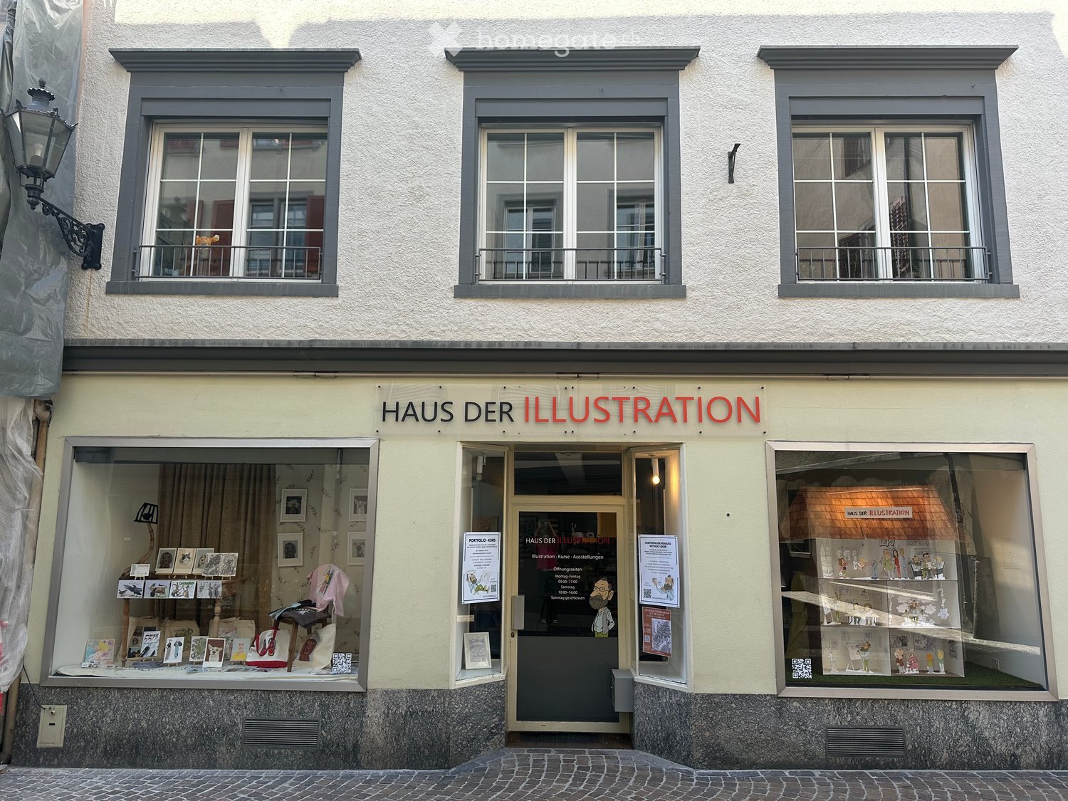 Facade of a building named "Haus der Illustration" with three windows and a central entrance. The windows display various framed artworks and illustrations.