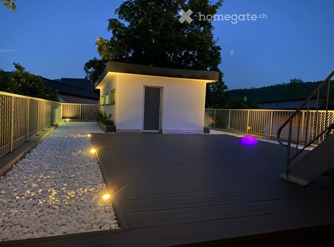 Modern outdoor deck with wooden flooring, integrated lighting, and a small white building with a grey door and windows.