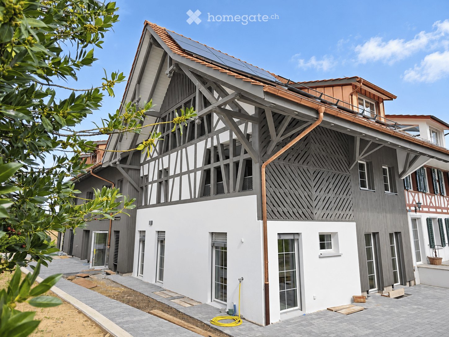 Modern two-story house with grey and white exterior, large windows, solar panels on the roof, and a balcony on the upper level.