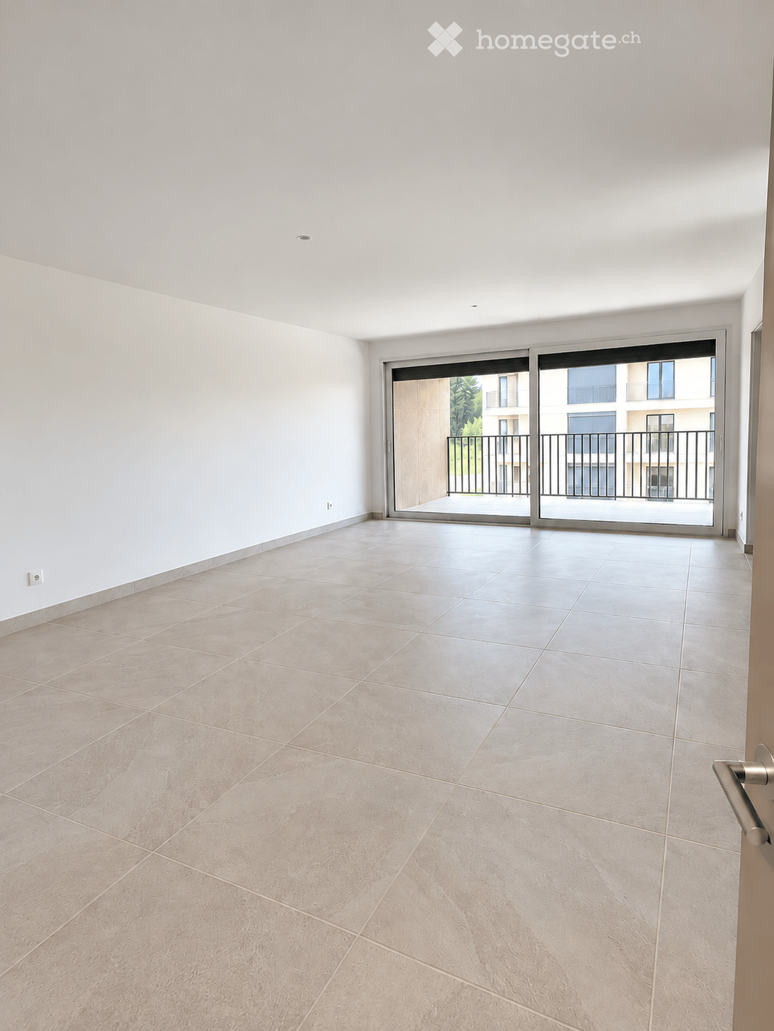 Empty room with light-colored tiled flooring and white walls, featuring a large sliding glass door leading to a balcony.