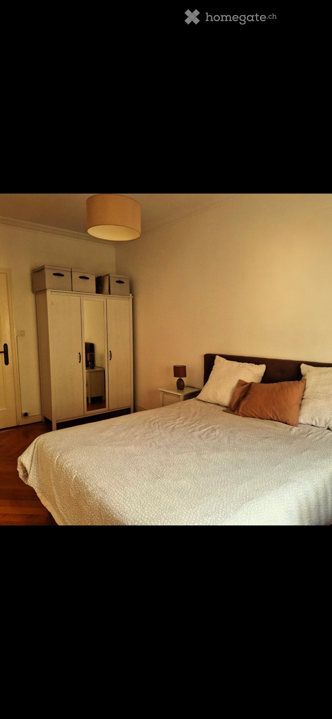 A well-lit bedroom with a large bed, nightstand, lamp, wardrobe, and mirror. The room features wooden flooring and neutral-colored walls.