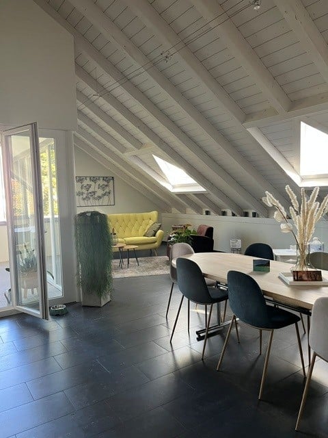 Bright, open-plan living and dining area with exposed wooden beams, skylights, dark flooring, a mustard-yellow sofa, a wooden dining table with modern chairs, and a tall decorative plant in the corner.