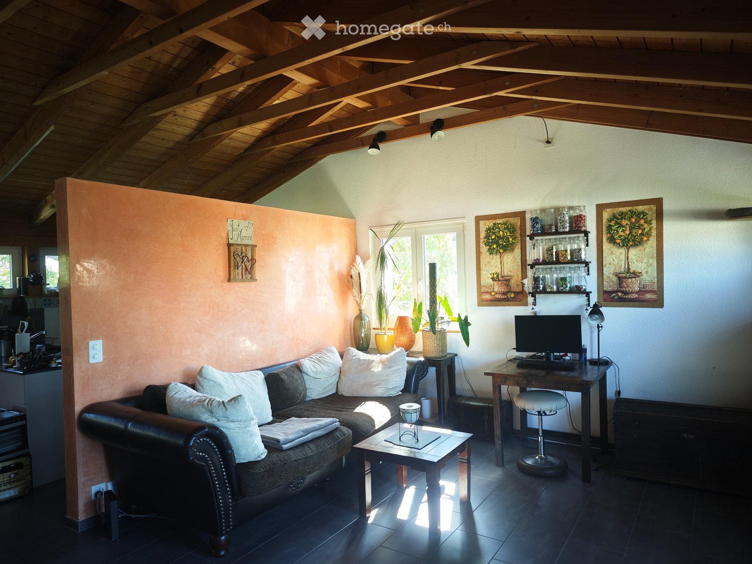 Cozy living room with exposed wooden ceiling beams, dark tiled floor, leather sofa with white cushions, coffee table, desk with computer, and decorative vases.