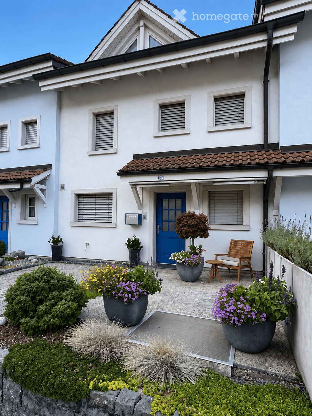 Front garden of a two-story house with a blue front door, potted plants, a wooden chair, and a small table.