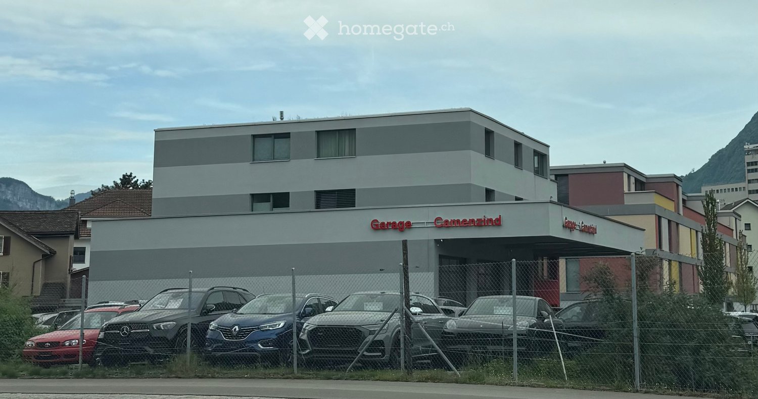 Modern multi-story building with a sign reading "Garage Camenzind" and a fenced parking area with several cars.