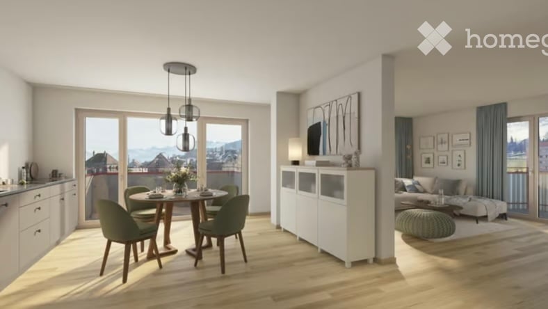 Modern, well-lit living and dining area with large windows offering a scenic view. Features a sofa, coffee table, dining table with chairs, a white cabinet, wall-mounted TV, floor lamp, and decorative picture frames.