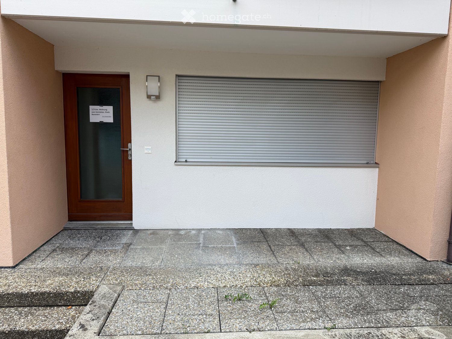 Entrance to a building with a wooden door, a closed metal shutter, and a light fixture above the door.