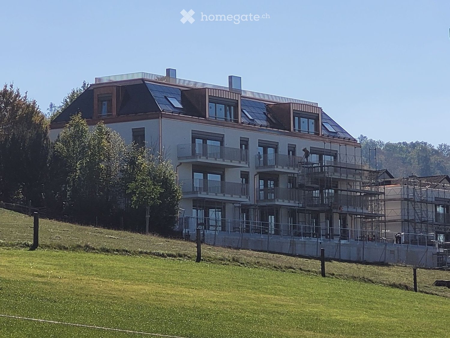 Modern multi-story building with balconies and a sloped roof, surrounded by greenery.