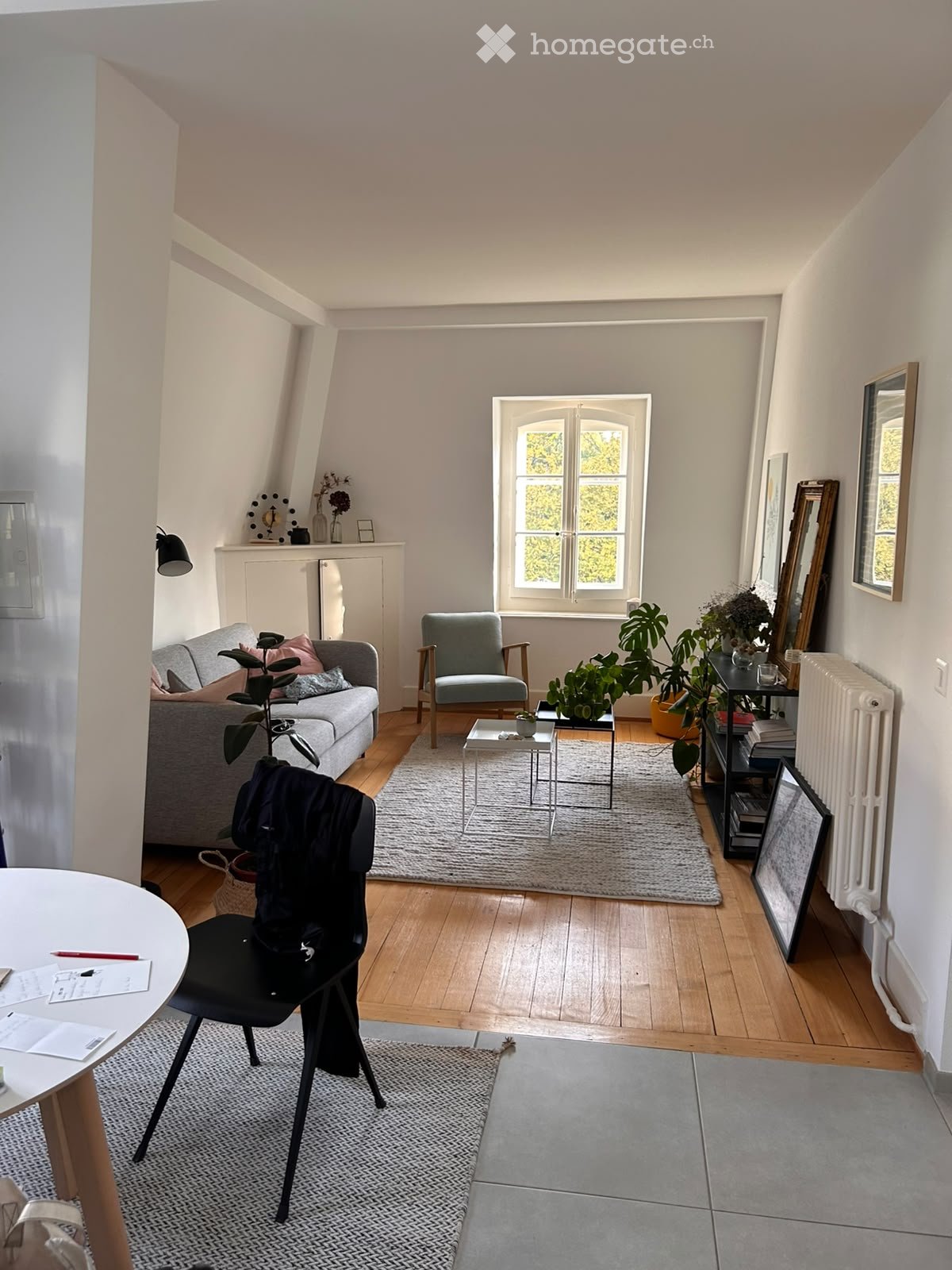 Bright living room with wooden flooring, large window, grey sofa, armchair, coffee table, and several plants. Includes a small dining area with a round table and chair.