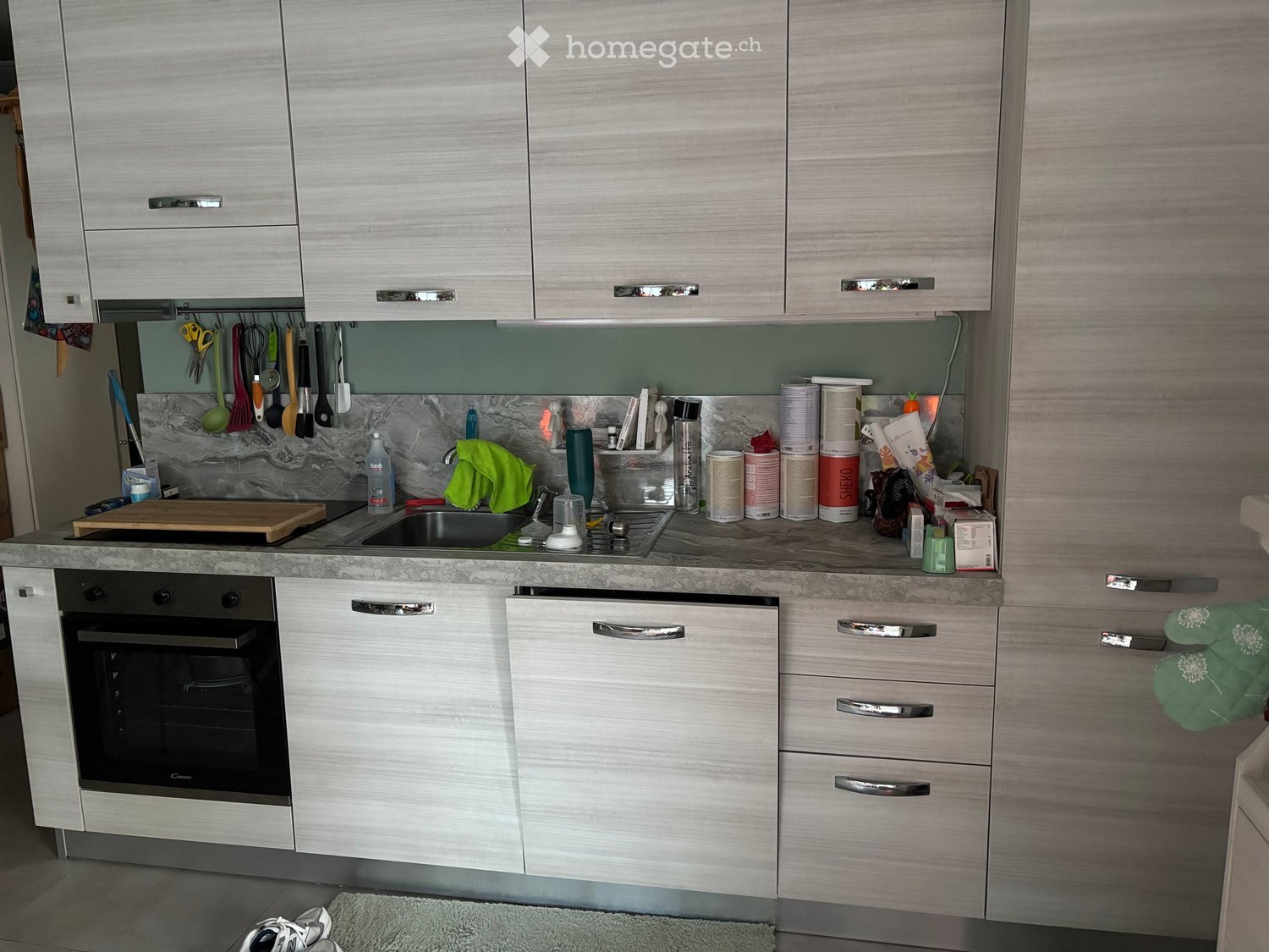 Modern kitchen with light wood cabinets, stainless steel appliances, marble backsplash, and various kitchen utensils and items on the counter.
