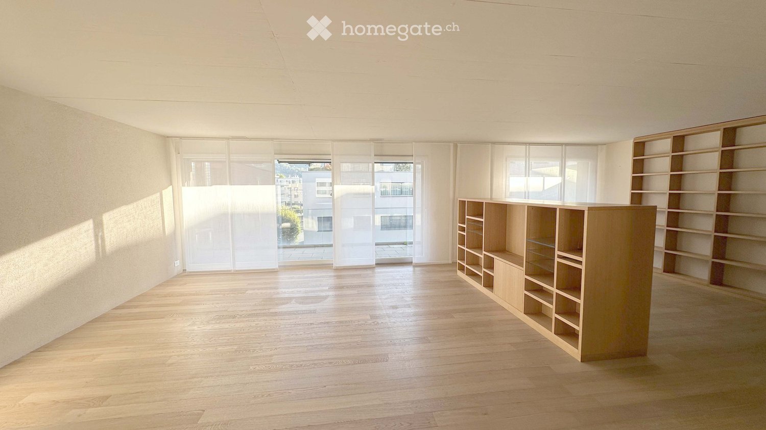 Empty room with wooden flooring, white walls, large windows with blinds, and built-in wooden shelving units.