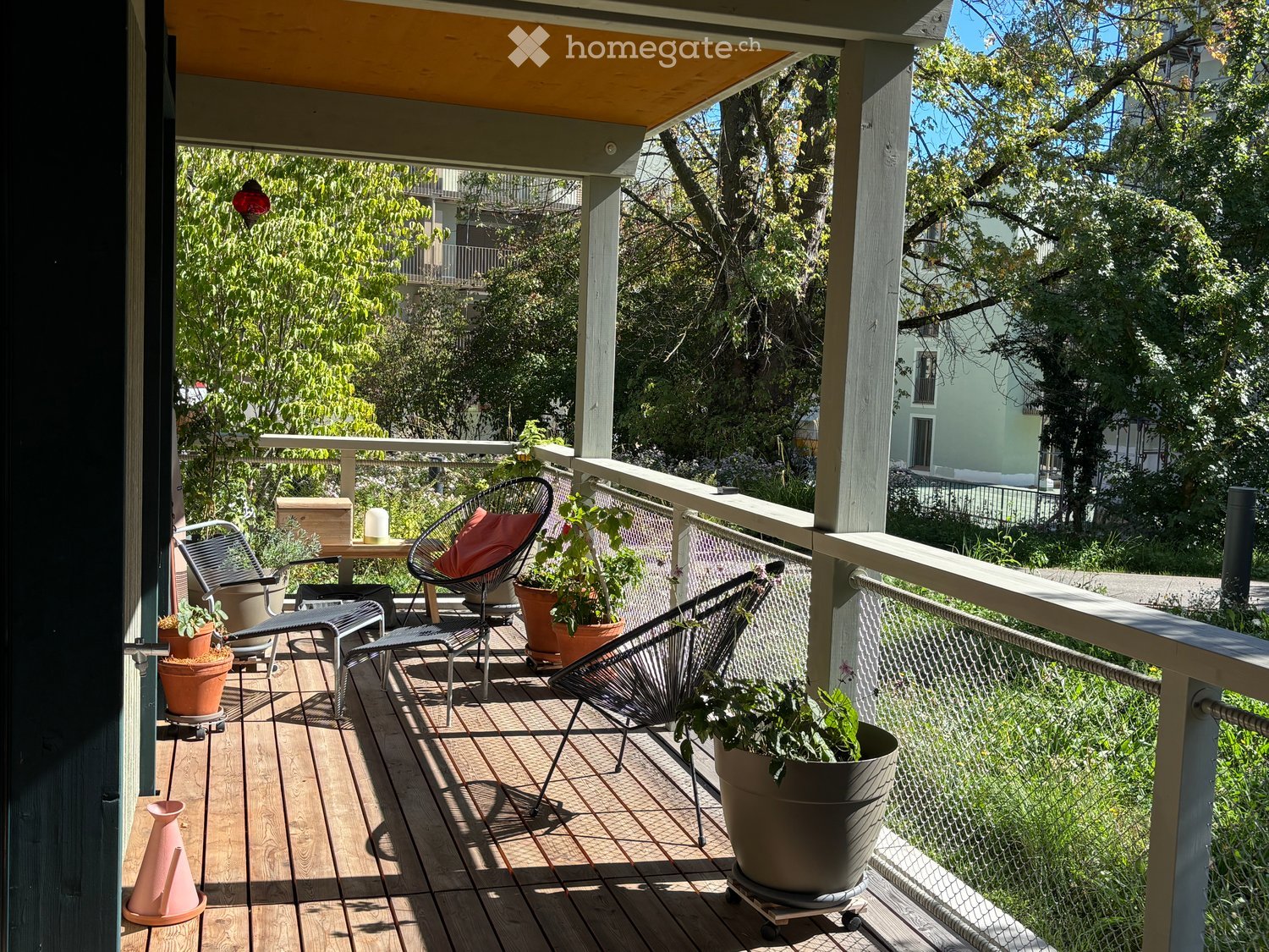 Balcony with wooden decking, metal railing, several chairs, a small table, and potted plants.
