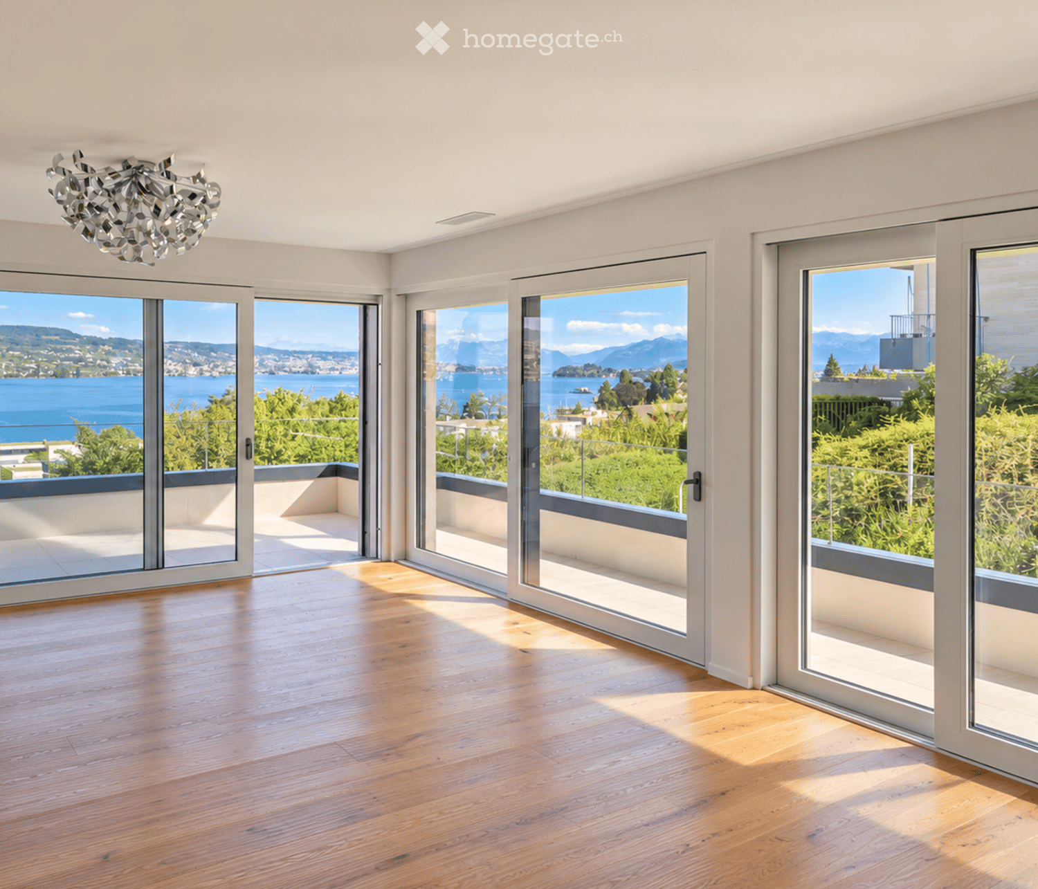 Bright, empty room with large glass sliding doors opening to a balcony, wooden floor, and a view of a lake and mountains.