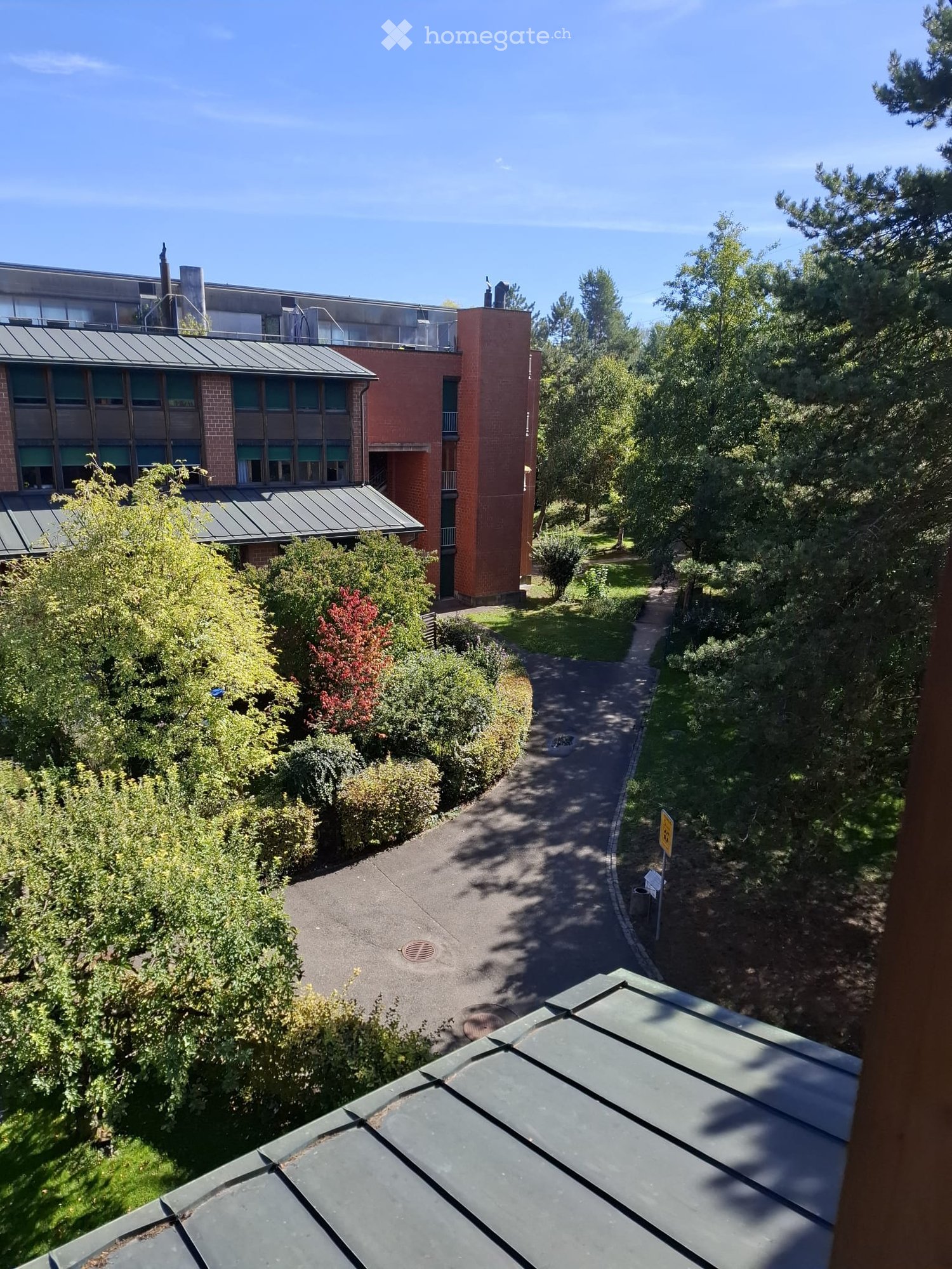 A modern multi-story building with red brick and grey metal roofing, surrounded by greenery and pathways.