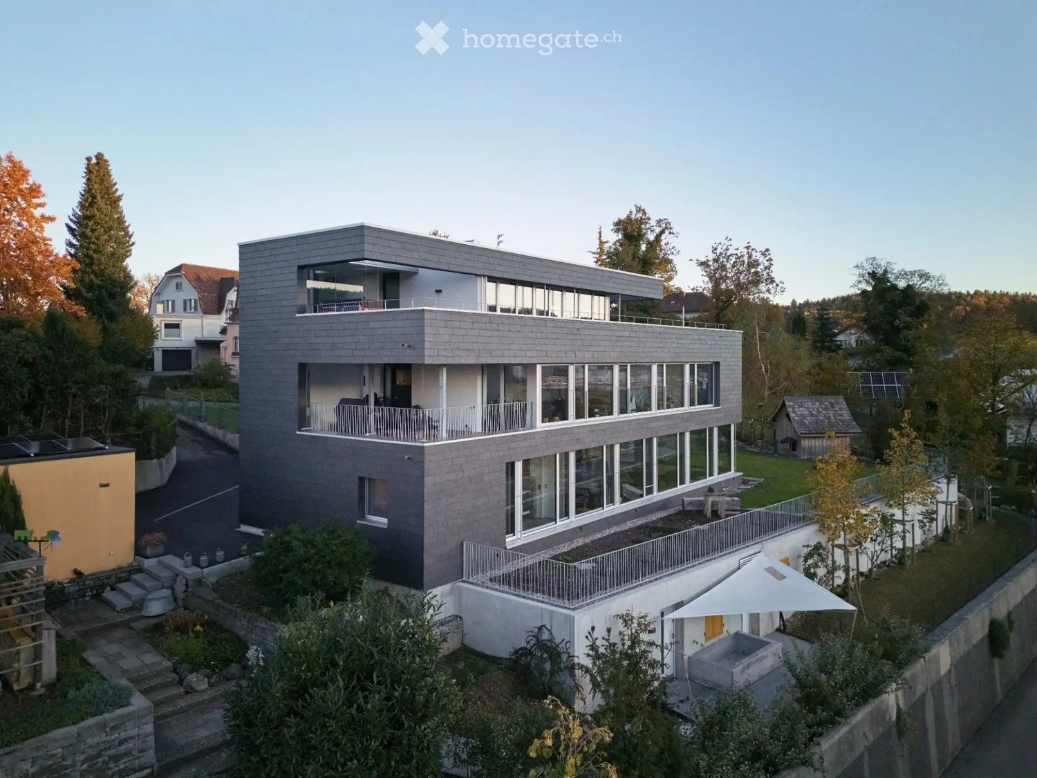 Modern two-story house with grey facade, large windows, balconies, and surrounding greenery.