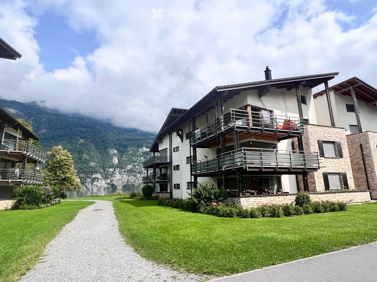 A modern two-story house with balconies, surrounded by a well-maintained lawn and garden, with a scenic mountain view in the background.