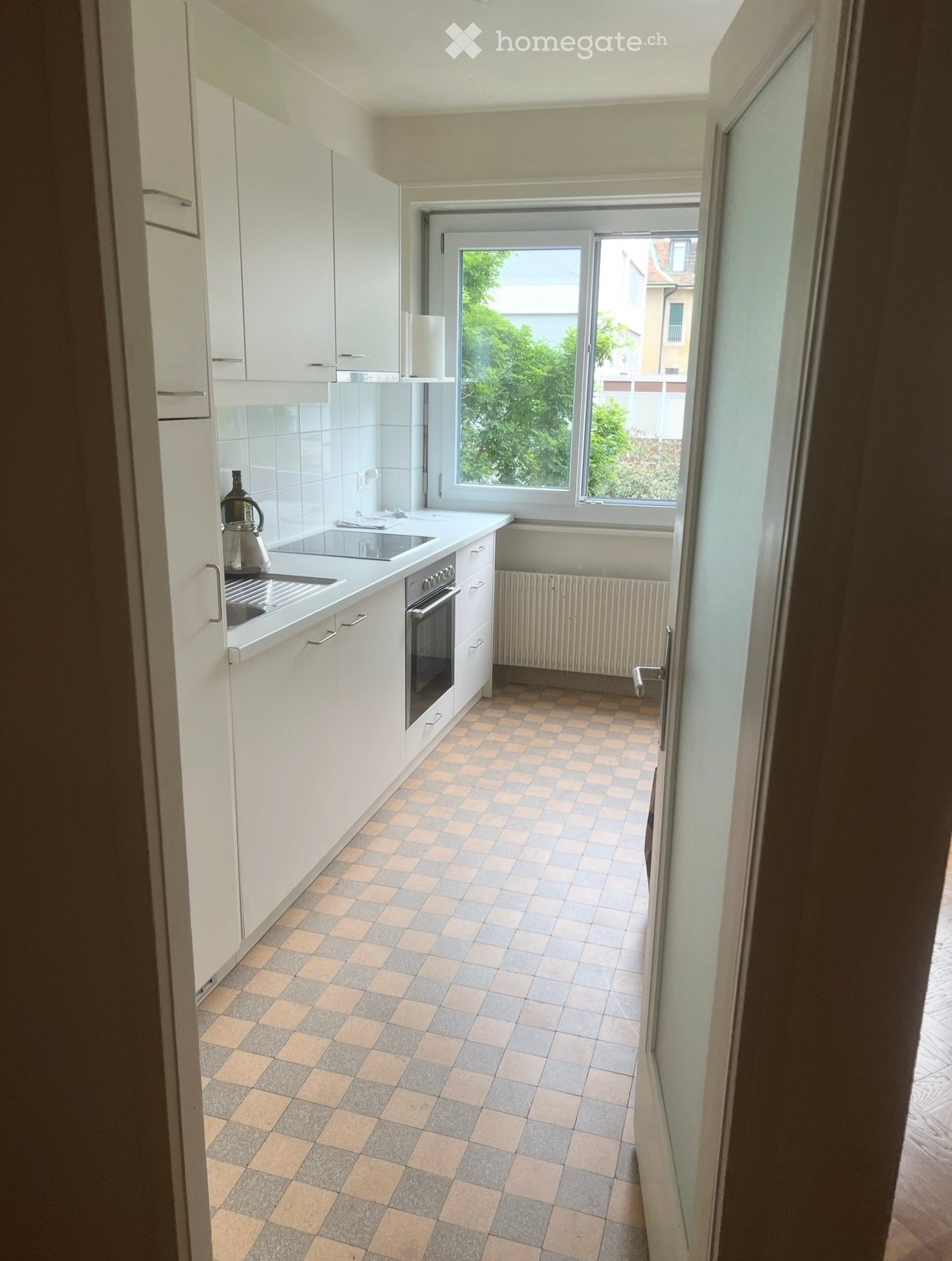 Modern kitchen with white cabinets, a tiled floor, and a window overlooking greenery. Includes a sink, oven, and teapot on the counter.