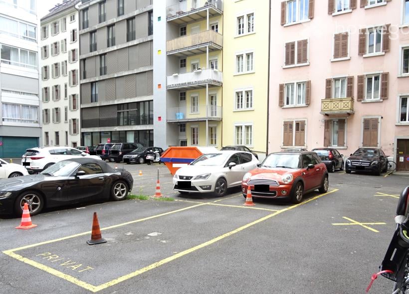 Urban parking lot with several cars, traffic cones, and surrounding multi-story apartment buildings with balconies and varied facades.