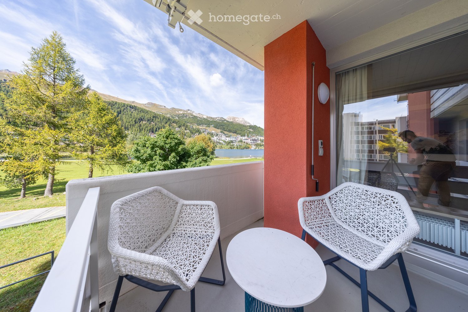 Balcony with two white wicker chairs and a round table, overlooking a scenic mountain view with trees and a lake.