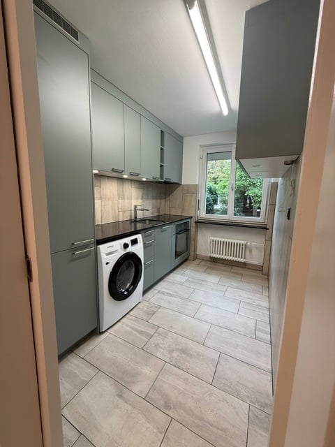 Modern kitchen with grey cabinets, a white washing machine, a sink with a faucet, and an oven. The floor is tiled, and there is a window with a view of greenery.