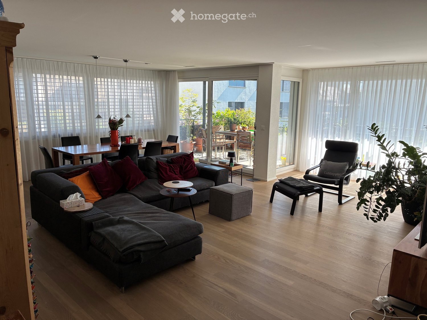 Modern living room with wooden flooring, large windows, and a balcony. Features a grey sectional sofa with colorful cushions, a coffee table, an ottoman, a dining table with chairs, and a potted plant near the window.