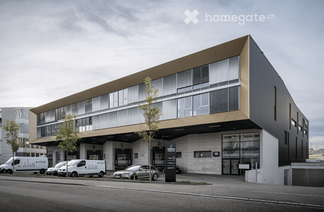 Modern multi-story building with large windows and a sleek design, featuring a parking area with several vans and a car.
