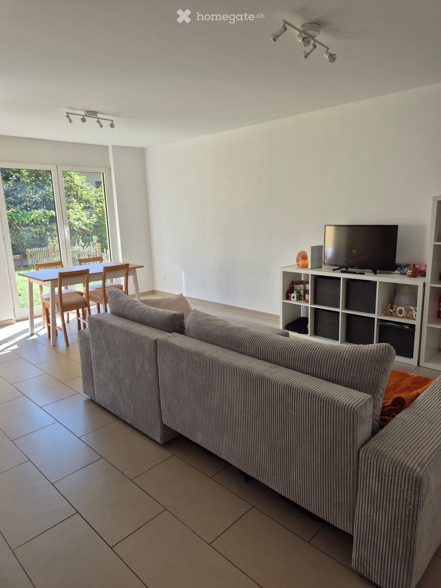 Modern living room with a grey sofa, a dining table with chairs, a television on a shelf, and large windows leading to a garden.