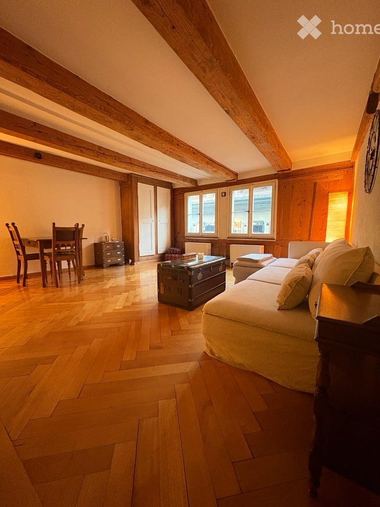 Cozy attic living room with wooden beams, hardwood floor, large windows, sofa, coffee table, dining area with table and chairs, and a small cabinet.