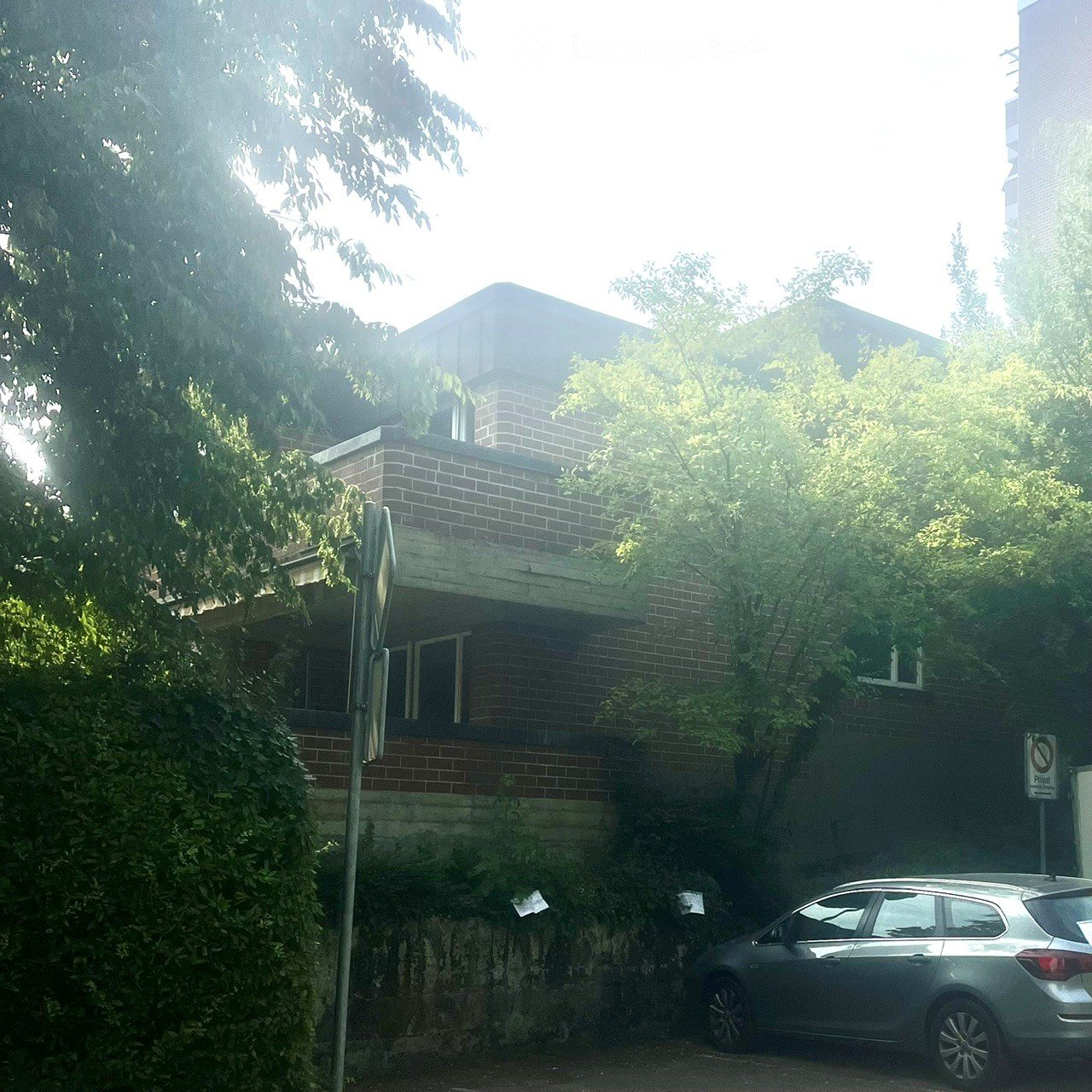 A two-story brick building with a flat roof and balcony, partially obscured by trees and foliage, with a parked car in the foreground.