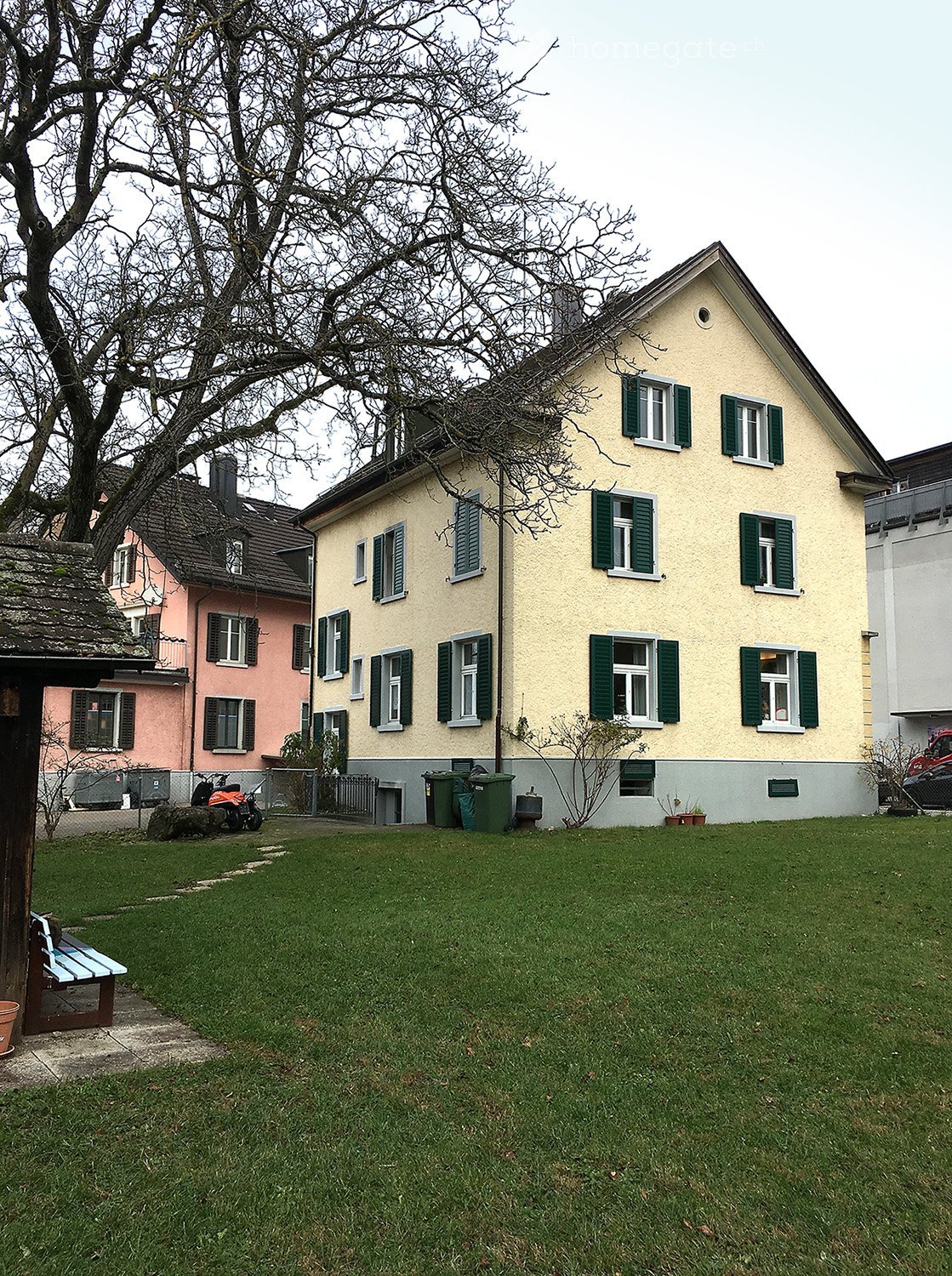 Lawn area in front of a two-story house with green shutters, a bench, and trash bins nearby.