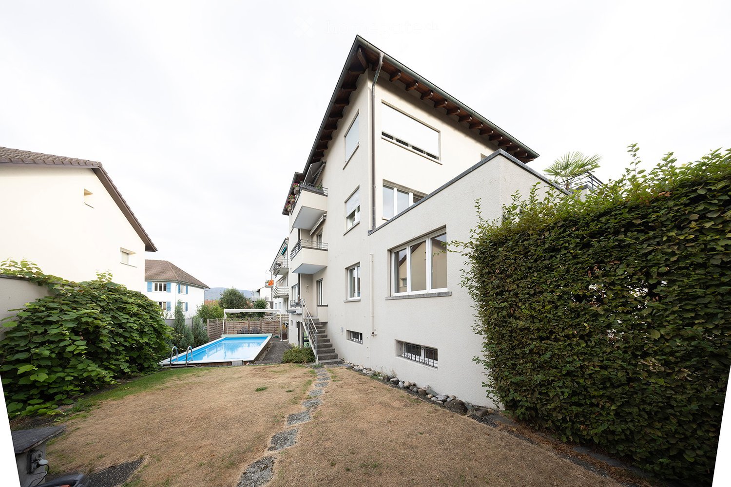Modern multi-story building with white exterior, large windows, balconies, and a swimming pool in the garden area.