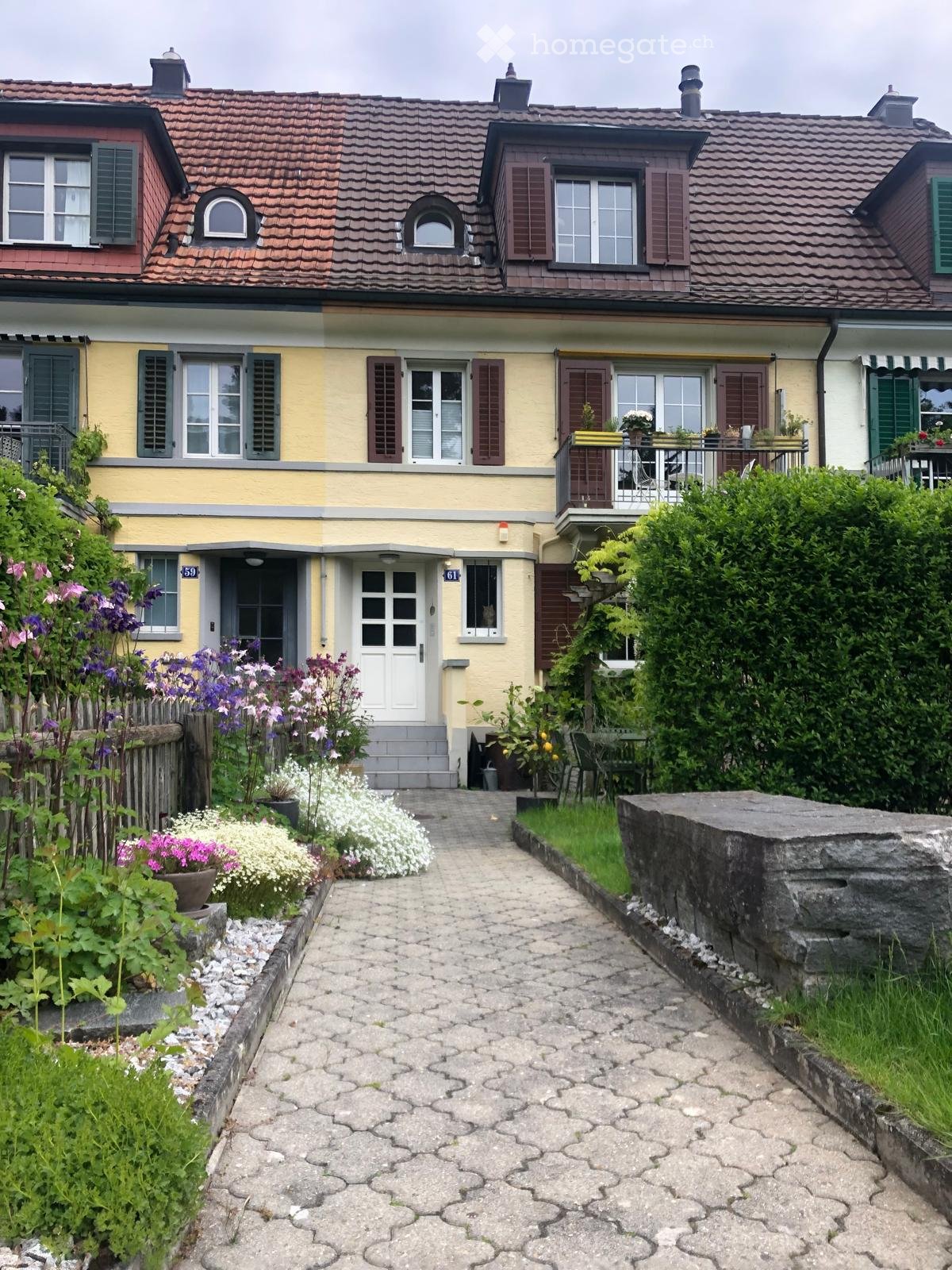 Two-story yellow house with red-tiled roof, multiple windows with shutters, and a small balcony. The entrance features a white door with a small porch, surrounded by a garden with flowers and a stone pathway.