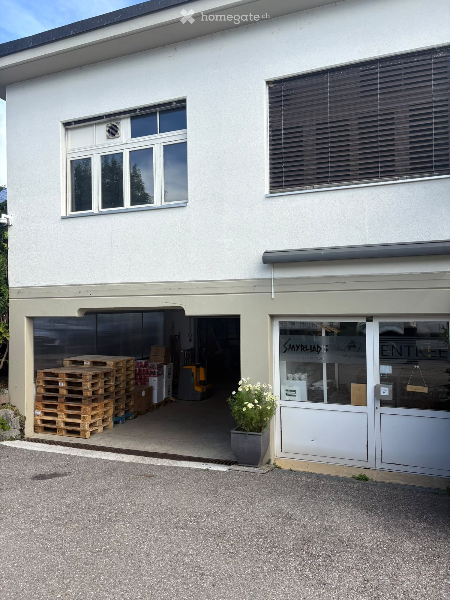 Two-story building with a garage entrance, stacked pallets, and a potted plant near the entrance.