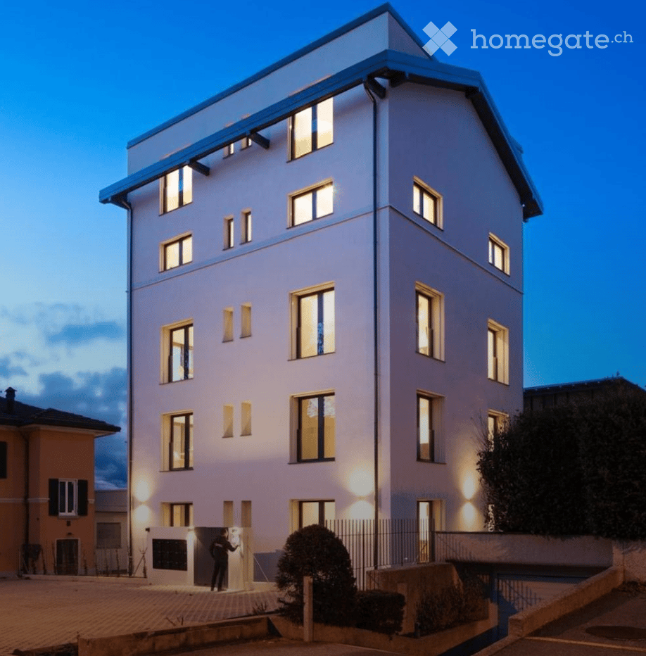 Modern multi-story residential building with illuminated windows and clean lines, featuring a raised ground floor and contemporary design.