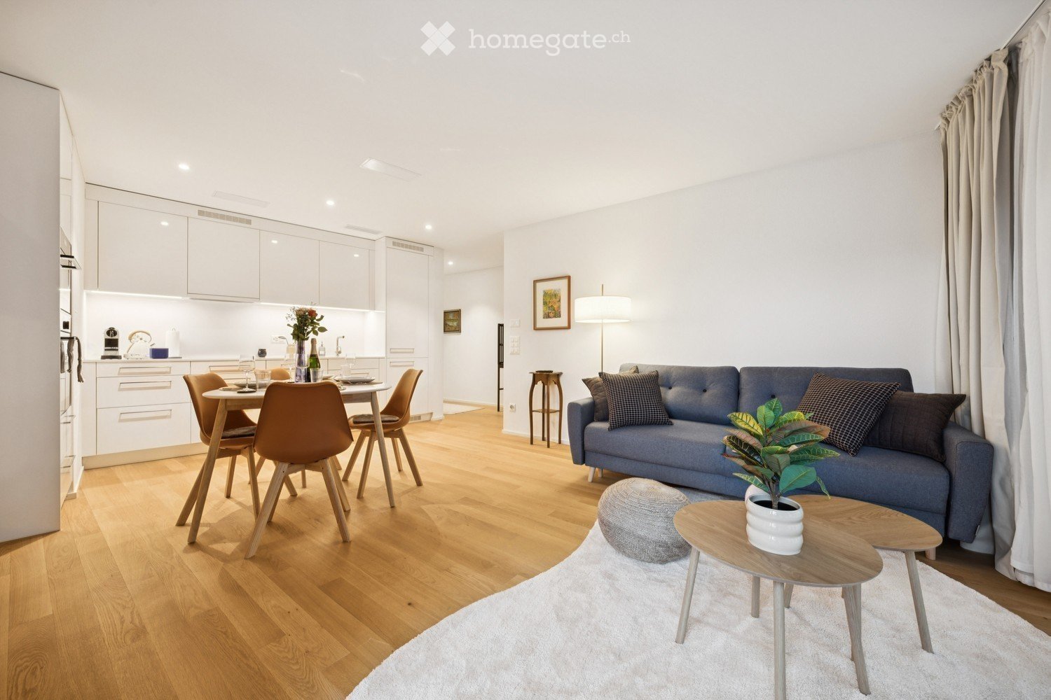 Modern open-plan living and dining area with light wood flooring, white walls, and large windows. Features a blue sofa with patterned cushions, a small coffee table with a potted plant, a grey ottoman, a dining table with four chairs, and a fitted kitchen