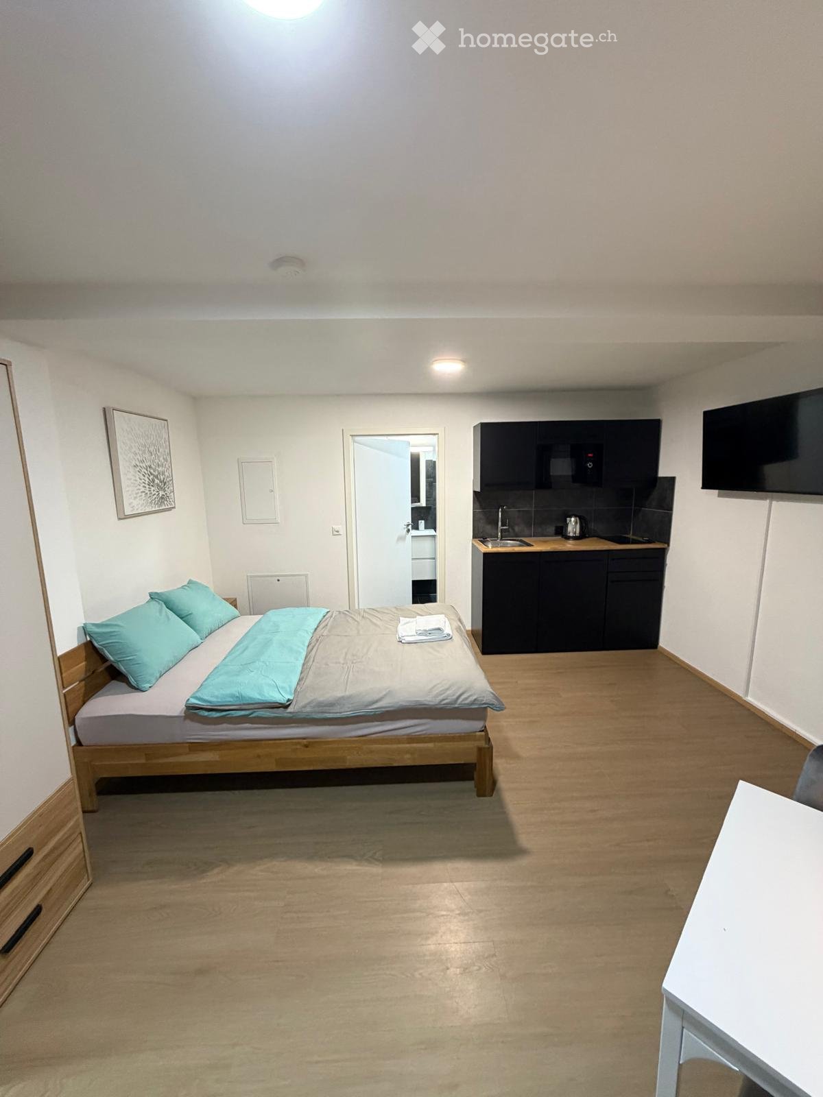 A small, furnished room with a bed, a table, a small kitchen area, and a TV screen mounted on the wall.