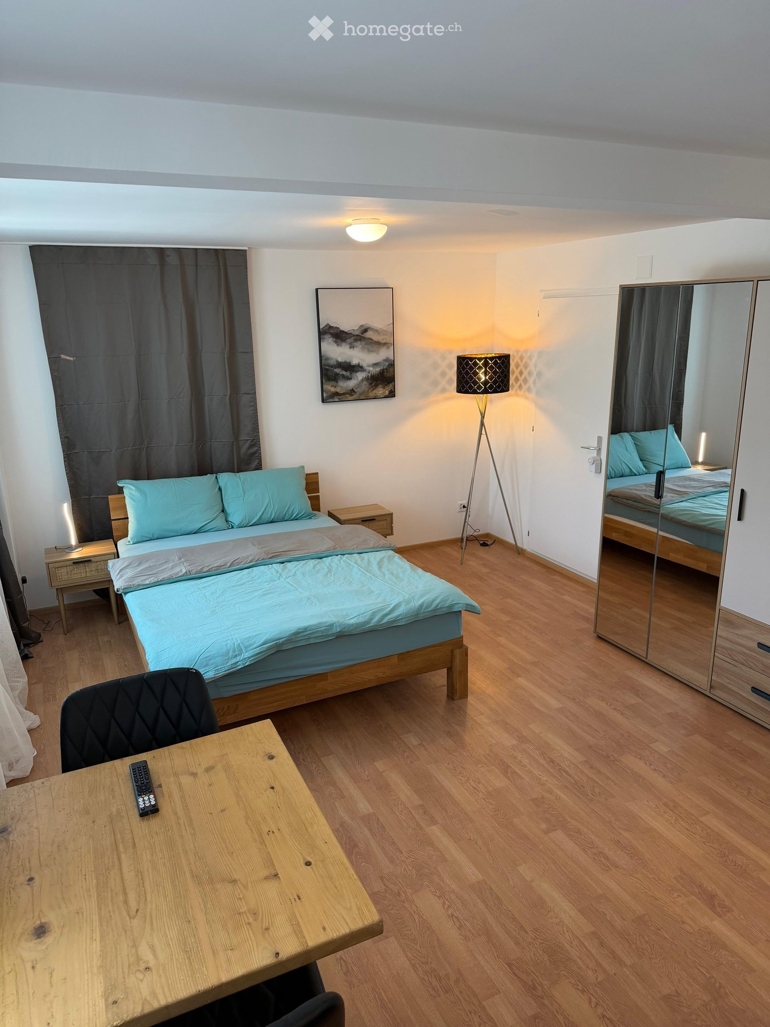 Cozy bedroom with wooden flooring, a double bed with blue bedding, a nightstand, a chair, a small table, a floor lamp, and a large mirror wardrobe.