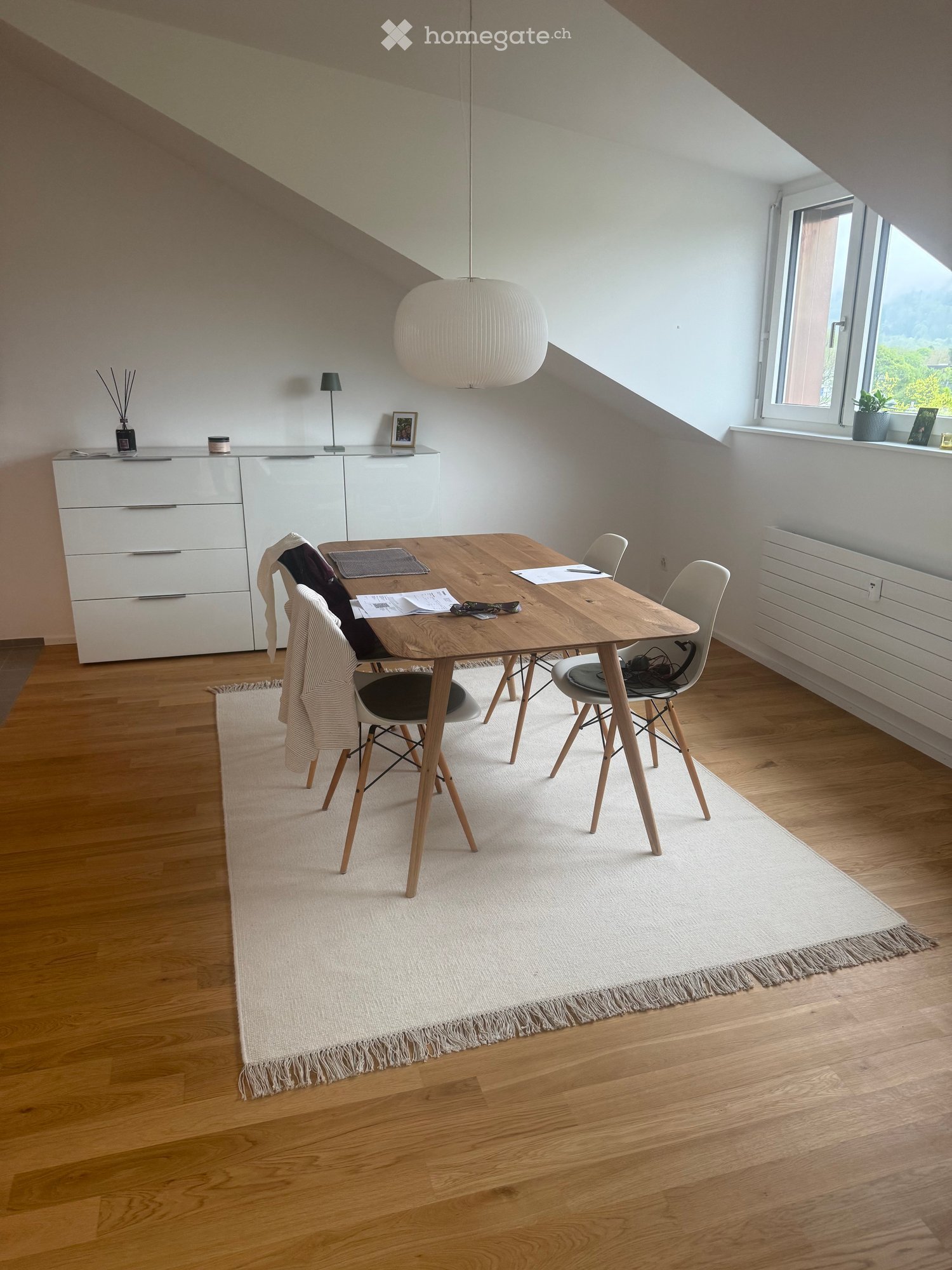 Bright, modern dining area with wooden table, four chairs, white cabinet, pendant light, and a large white rug.