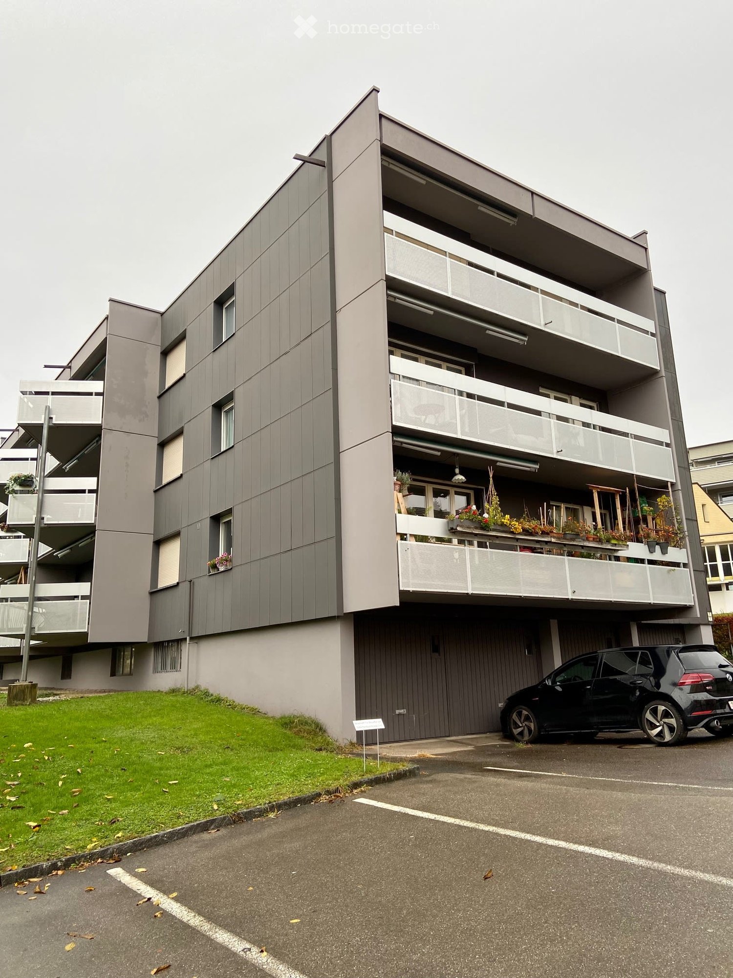 Modern multi-story apartment building with grey facade, balconies, and potted plants. A black car is parked in the driveway.