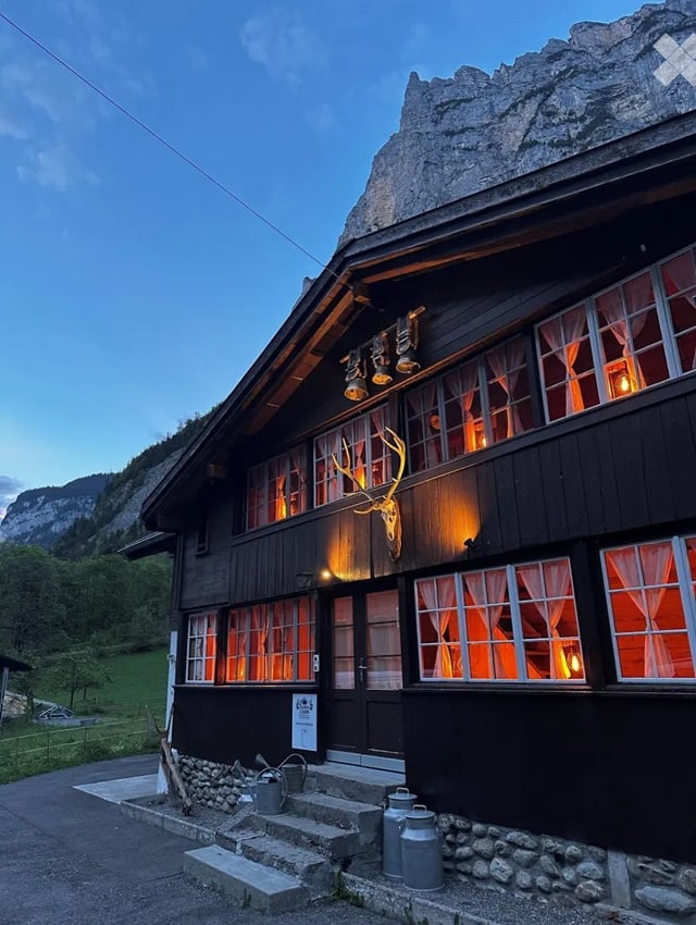 A rustic wooden building with large windows, warm interior lighting, and a mounted deer head on the facade. The building is surrounded by a stone pathway and has a mountainous backdrop.