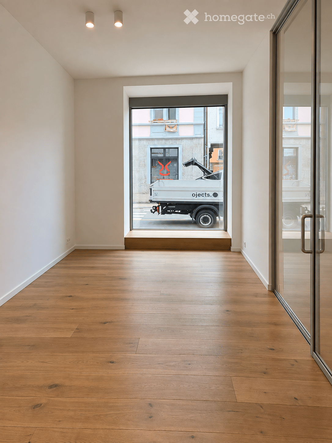 Empty room with wooden flooring, white walls, and large glass windows showing a street view.