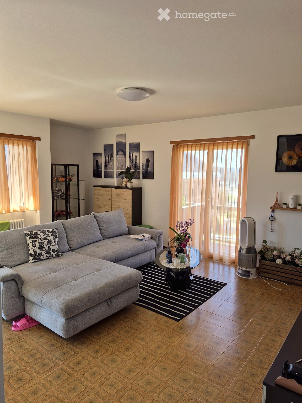 Modern living room with grey sectional sofa, coffee table, fan, and decorative items. Features large windows with curtains, wooden flooring, and artwork on the walls.