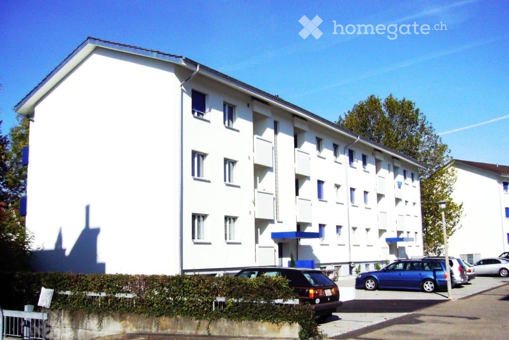 A modern, multi-story residential building with white walls and blue accents, featuring multiple windows and a parking area with several cars.